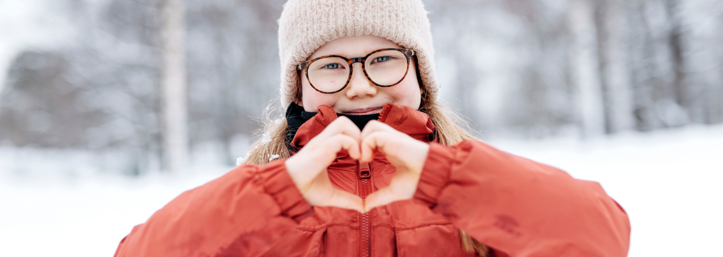 Hymyilevä ihminen talvivaatteissa näyttää käsillään sydämen merkkiä ulkona talvisessa säässä.