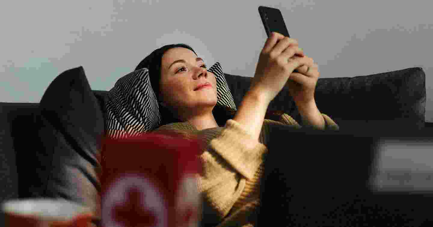 Hymyilevä henkilö makaa selällään sohvalla, kädessään älypuhelin.