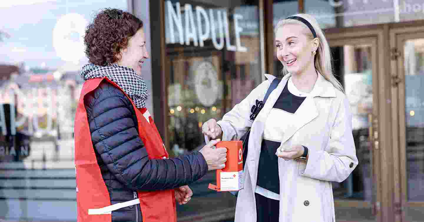 Punaisen Ristin vapaaehtoinen lipaskerääjä ottaa hymyilevältä henkilöltä vastaan lahjoituksen keräyslippaaseen.