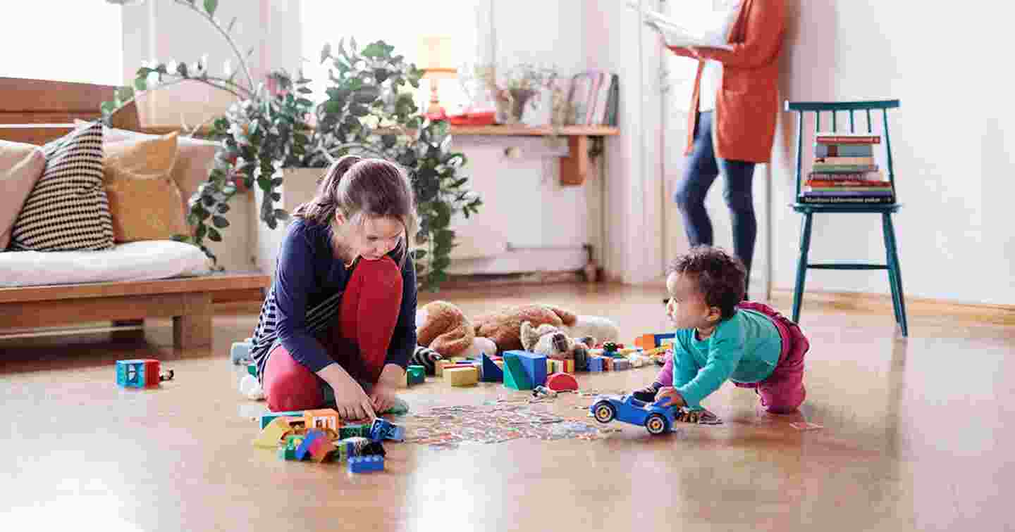 Kaksi lasta leikkii asunnon lattialla leluillaan.