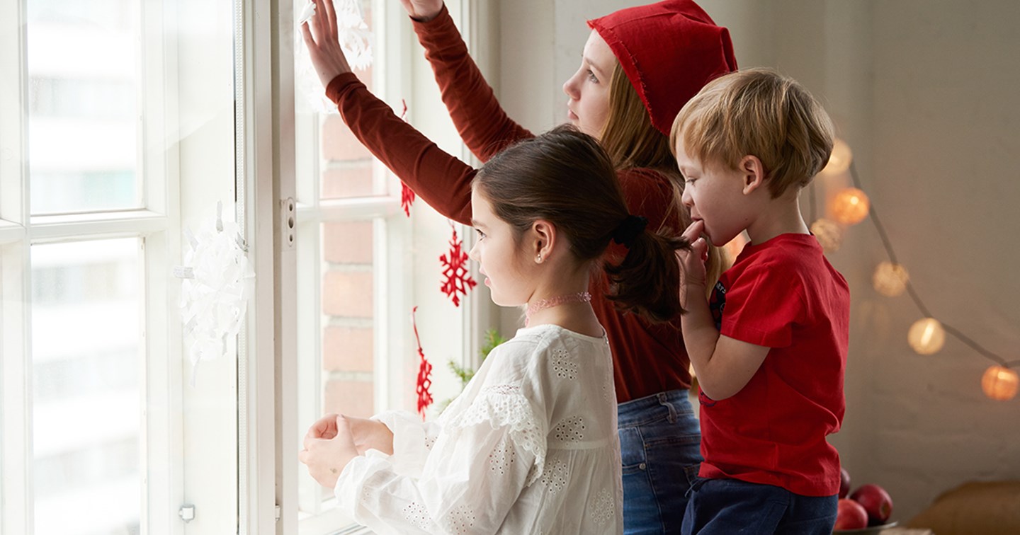 Kolme lasta jouluisissa vaatteissa katsoo ulos ikkunasta.