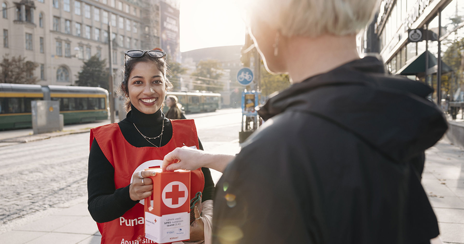 Hymyilevä lipaskerääjä Suomen Punaisen Ristin liiveissä pitelee lipasta, johon ohikulkija laittaa kolikon.