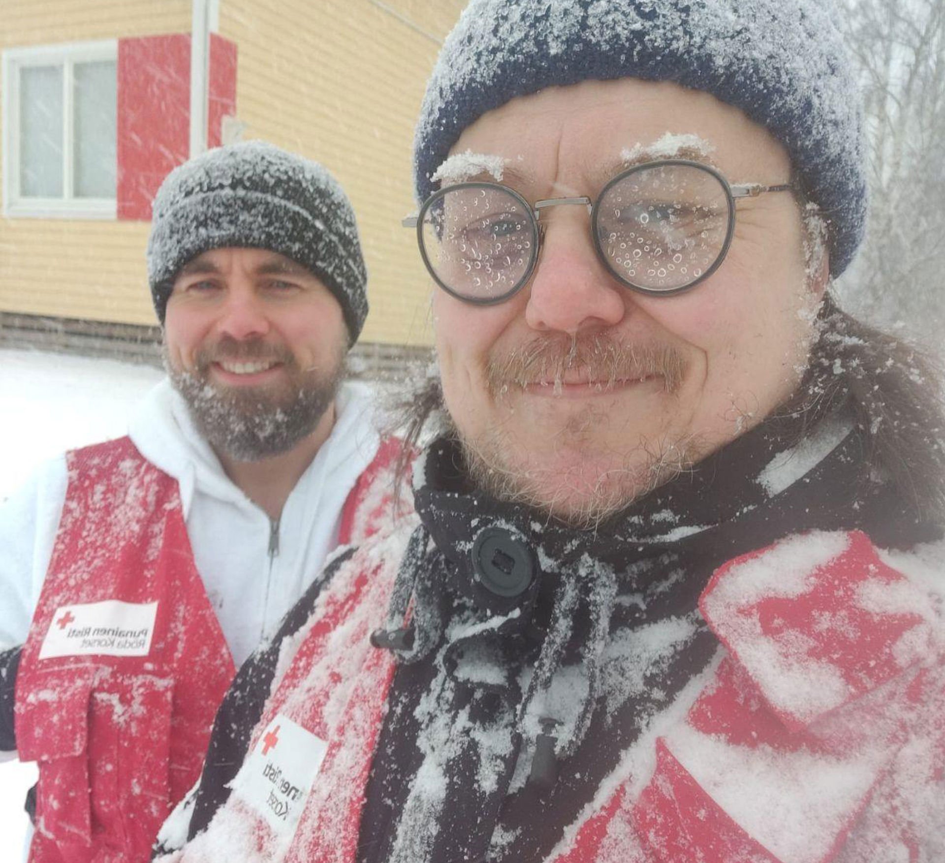 Teemu Hiilinen ja Vesa Vahermaa seisovat hymyillen hyvin lumisessa säässä yllään Punaisen Ristin vapaaehtoisliivit.