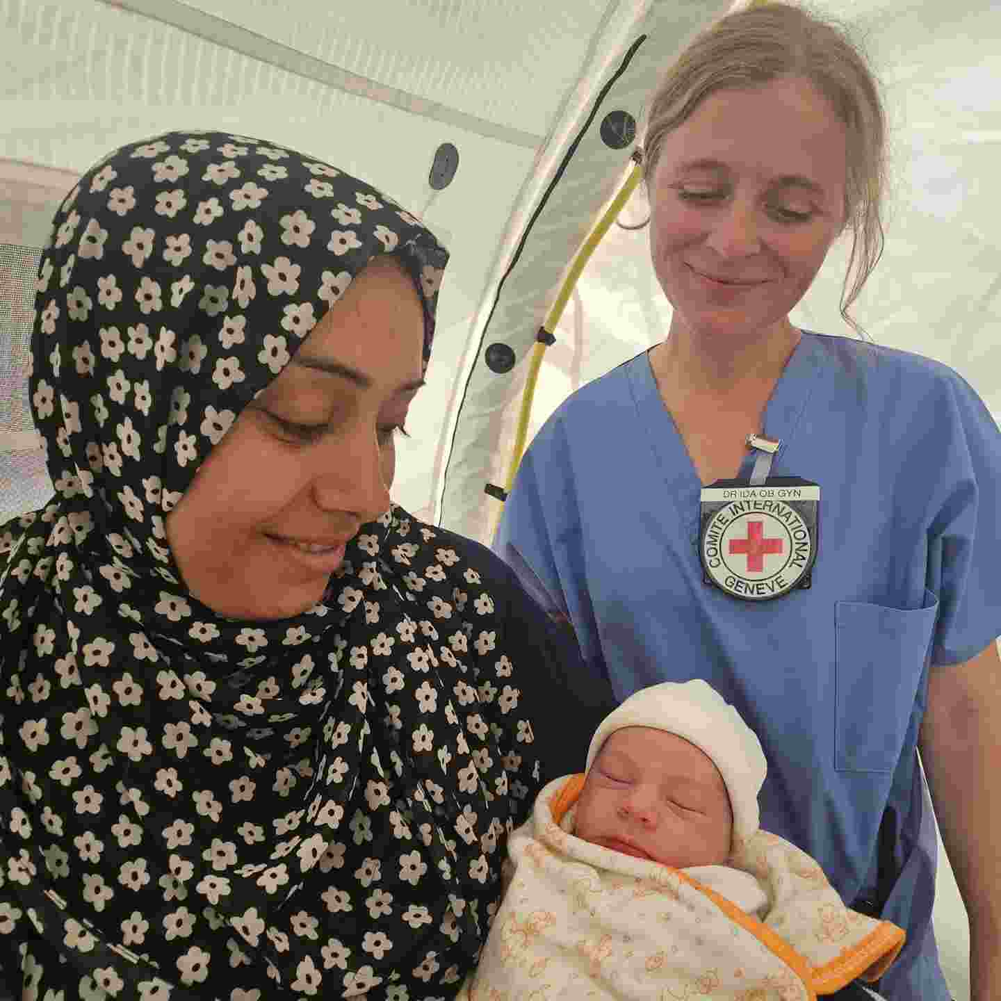 Hymyilevä nainen pitää sylissään vastasyntynytta vauvaa. Vieressä Punaisen Ristin hymyilevä työntekijä.
