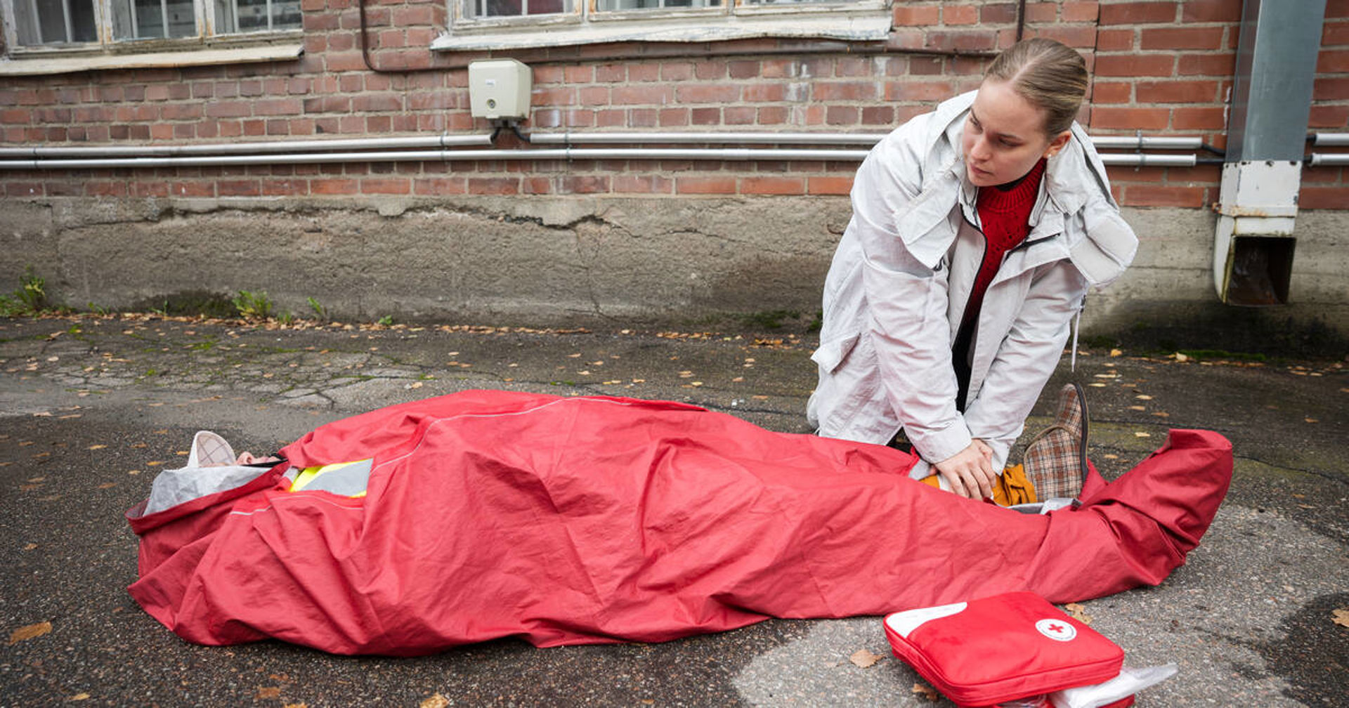 Henkilö makaa maassa punaisen lämpöpeitteen alla ja toinen henkilö on polvistunut hänen viereensä ja tarkistaa hänen tilaansa. Vieressä kuvassa näkyy ensiapulaukku. Tilanne tapahtuu ulkona tiilirakennuksen edustalla ja maassa on pudonneita lehtiä. 