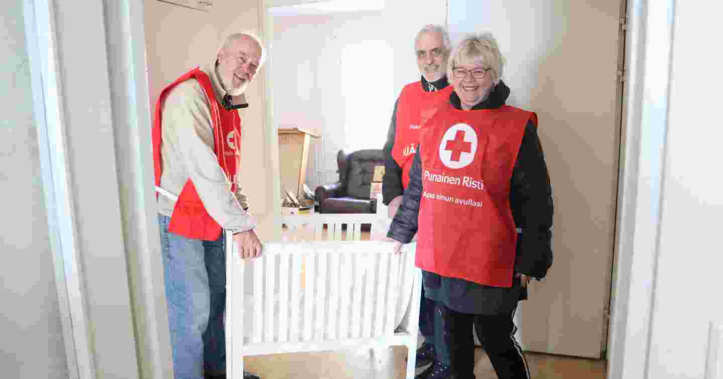 Kolme hymyilevää ihmistä Punaisen Ristin vapaaehtoisliiveissä seisovat kehdon vieressä pakolaisille kalustamassaan asunnossa