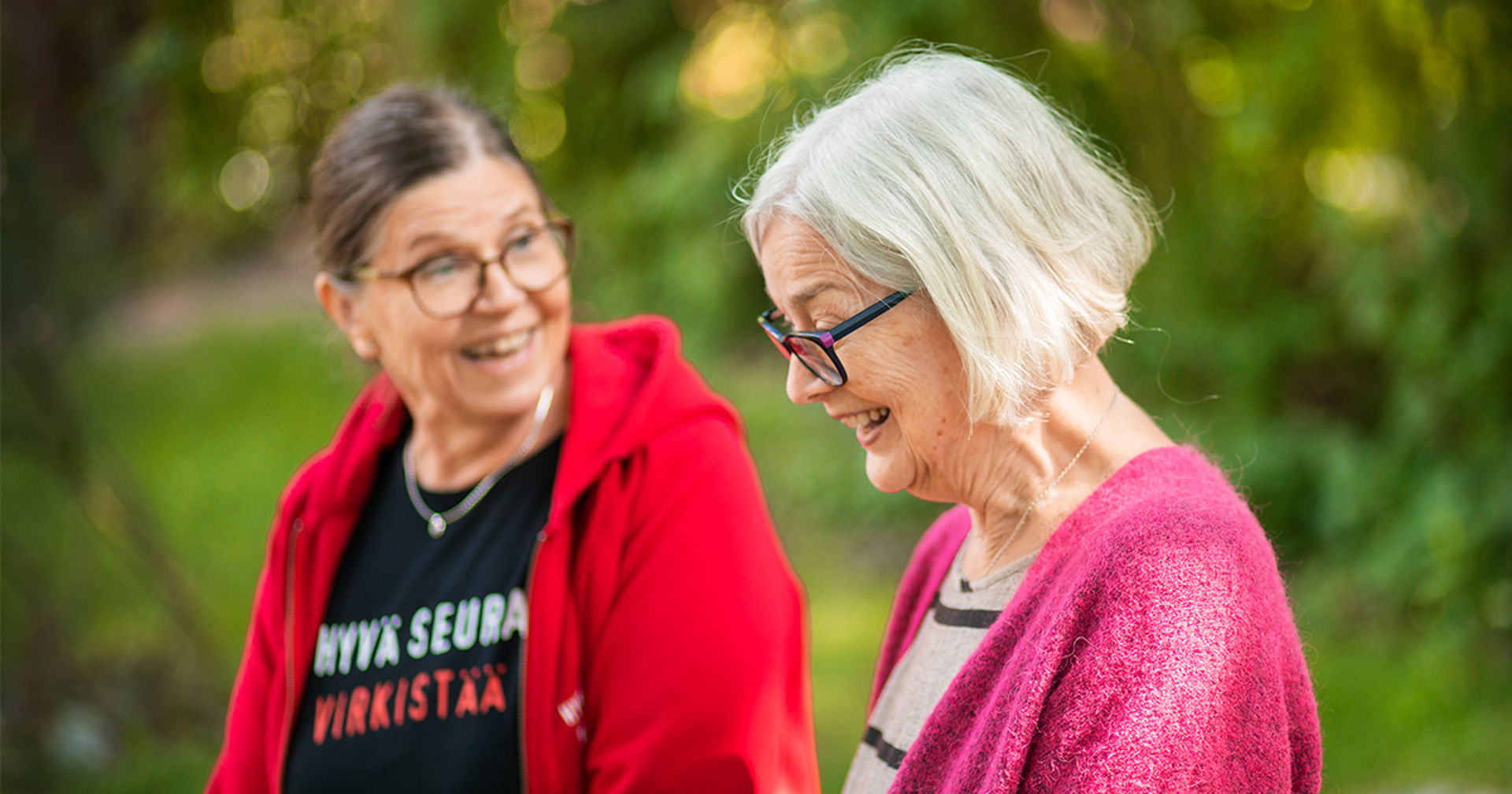 Kaksi henkilöä keskustelee hymyillen. Taustalla näkyy vehreää luontoa.