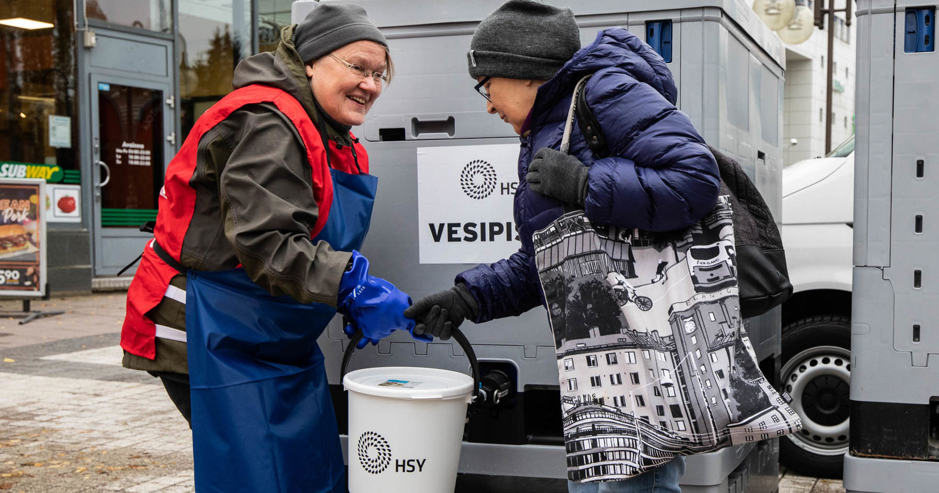 Hymyilevä henkilö Punaisen Ristin vapaaehtoisliiveissä ojentaa kannellista ämpäriä naiselle, jonka vedenjakelu on katkennut. Punainen Risti suorittaa vedenjakelua kotimaan kriisitilanteissa.