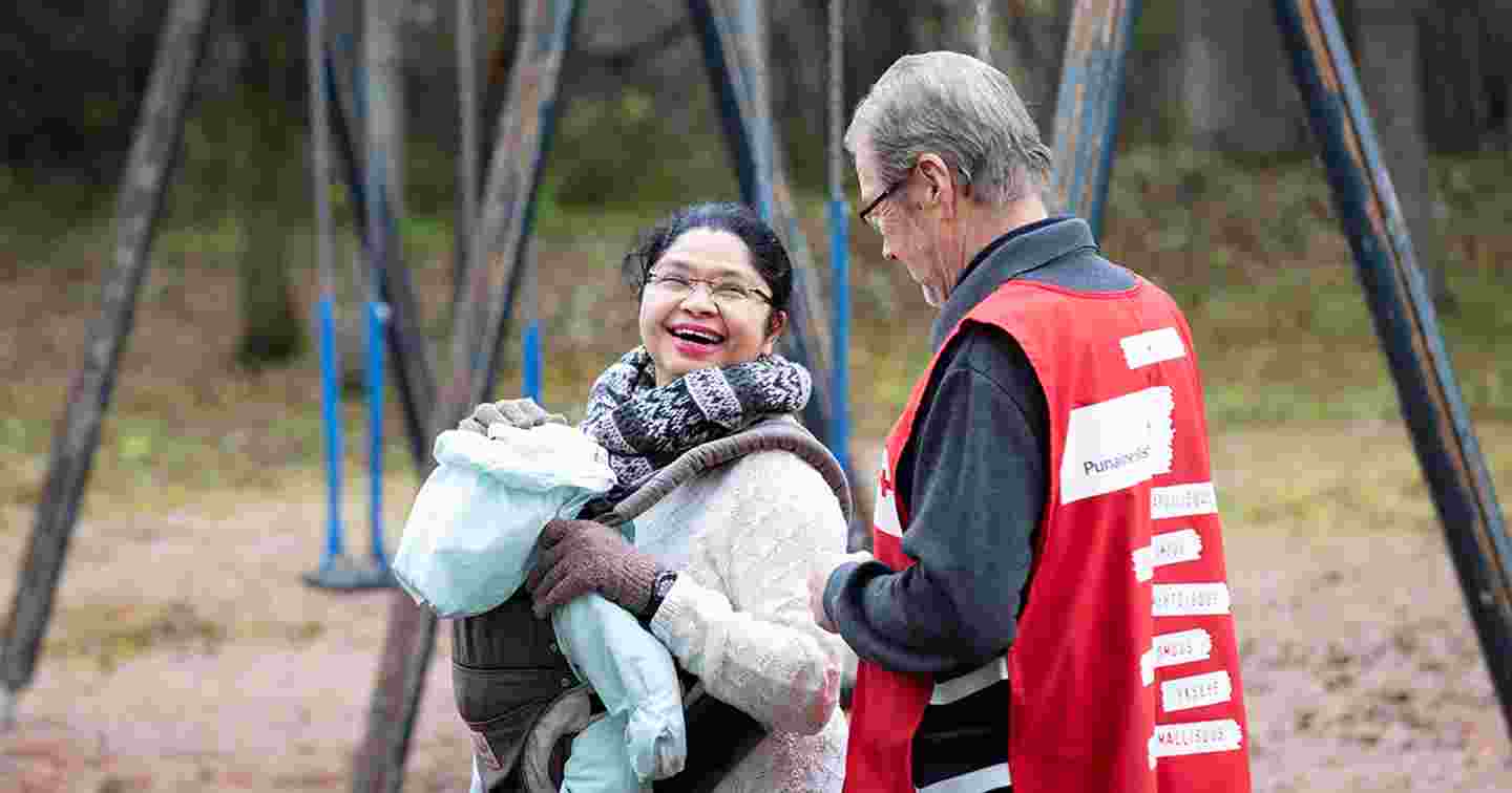 Henkilö Punaisen Ristin vapaaehtoisliiveissä opettaa iäkästä maahanmuuttajanaista ja -lasta. Iäkäs nainen halaa vapaaehtoista.