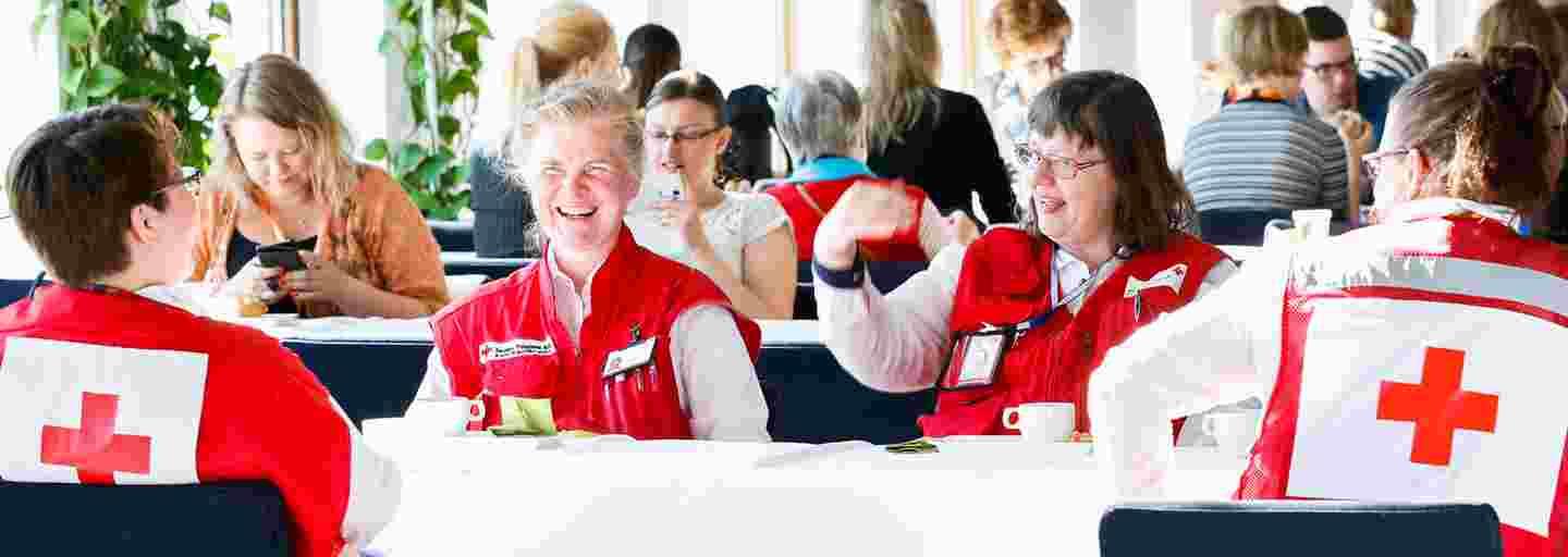 Neljä hymyilevää henkilöä Punaisen Ristin varusteissa istuu väkijoukossa Punaisen Ristin yleiskokouksessa Finlandia-talossa.