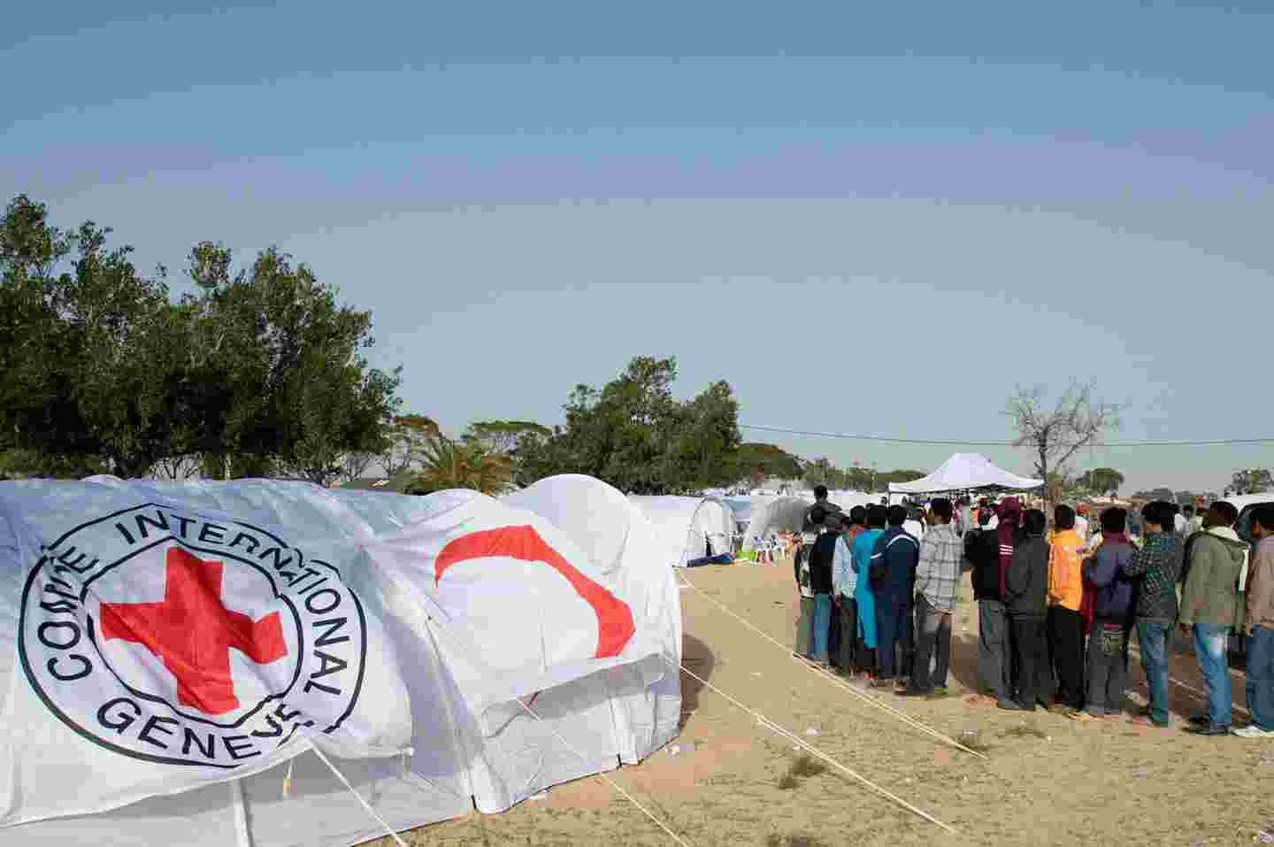 Ihmisiä jonossa pakolaisleirilla Punaisen Ristin tunnuksin varustettujen telttojen keskellä.