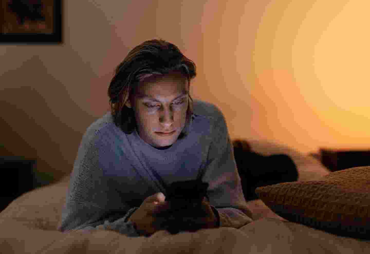 Nuori käyttää älypuhelinta sängyssään hämärässä valaistuksessa.